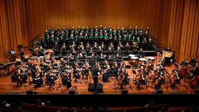 Large orchestra, piano and choir on stage with Louis Sharpe conducting.