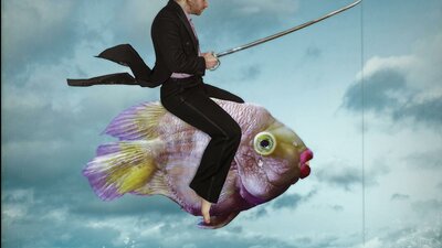 A man wearing a black suit riding a fish through the cloudy sky