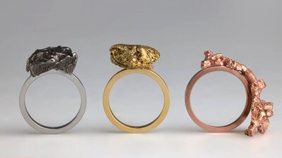 Three rings with raw lumps of metal stand upright; one silver, one gold and one copper