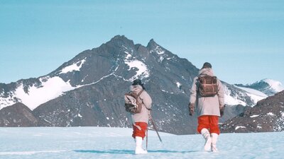 Two hikers walking on snowy ground, in front of snow-covered peaks.