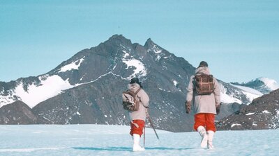 Two hikers walking on snowy ground, in front of snow-covered peaks.