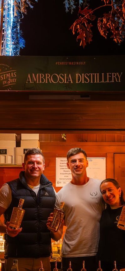 Ambrosia Distillery at Christmas in July Exhibitors standing in their wooden chalet decorated with Christmas lights, selling liquor