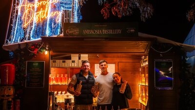 Ambrosia Distillery at Christmas in July Exhibitors standing in their wooden chalet decorated with Christmas lights, selling liquor