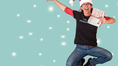 RA Spratt, wearing a santa hat and holding a book, leaps jubilantly in the air, as snow falls.