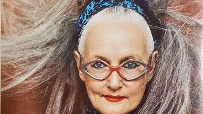 A woman with diamond-shaped classes smiles smartly. Her long grey ponytail sweeps around her.