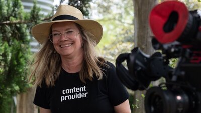 A blonde woman in a beige hat wears a black t-shirt. She stands smiling next to a camera