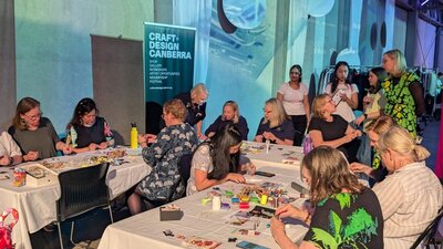 At a u-shaped table, a group of people sit and create small wove tokens out of recycled materials.