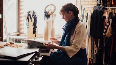 Kelli Donovan, Circular Fashion Designer A woman with a blue scarf sits in profile at a table, reading a book. Behind her are 2 mannequins.