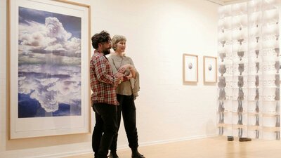 Two people viewing artwork in a bright contemporary gallery, with framed prints and an installation.