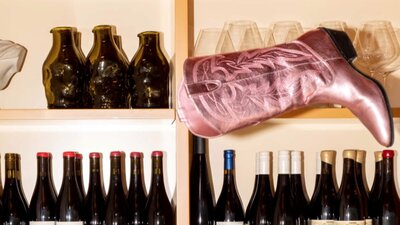 A metallic pink cowboy boot sits at a peculiar angle on a shelf full of wine