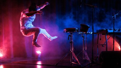 Man jumping on a pink and purple lit stage with percussion equipment