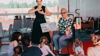 Community day in the Garden An Auslan interpreter standing next to a person reading a book to a group of children sitting down
