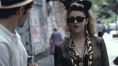 Woman walking towards man wearing 1980s style clothing, sunnies and big hair bow