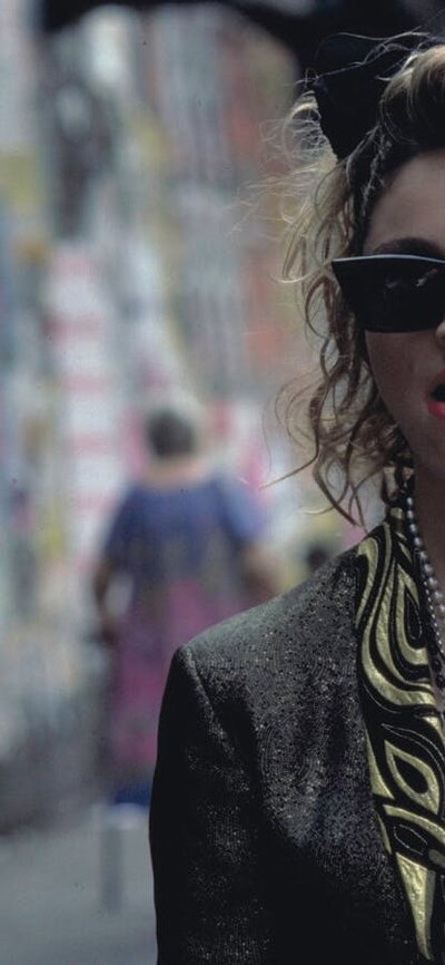 Woman walking towards man wearing 1980s style clothing, sunnies and big hair bow