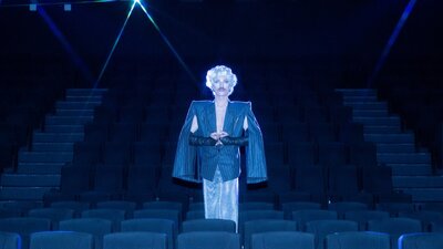 Venus Mantrap Drag artist stands in the middle of an empty cinema wearing an overcoat and blonde wig