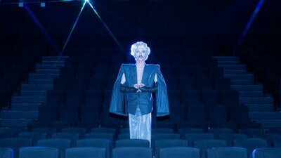 Venus Mantrap Drag artist stands in the middle of an empty cinema wearing an overcoat and blonde wig