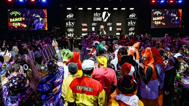 Fans in fancy dress fill the floor for a night of Premier League darts. A crowd of fans in colourful fancy dress standing in front of a brightly lit darts stage.
