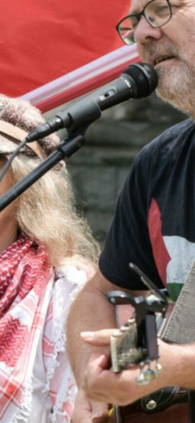 David Rovis & Kamala Emanuel singing live at a protest in front of a Palestinian flag.]