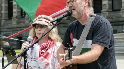 David Rovis & Kamala Emanuel singing live at a protest in front of a Palestinian flag.]
