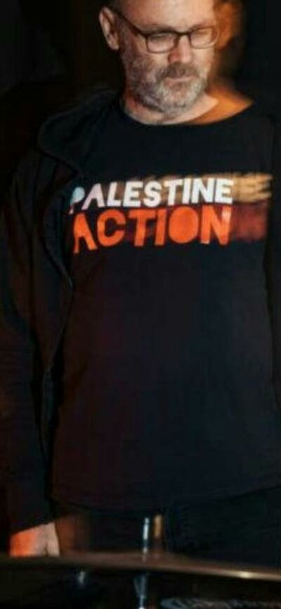 David Rovics standing behind drums with a Palestine Action t-shirt on. Black background.