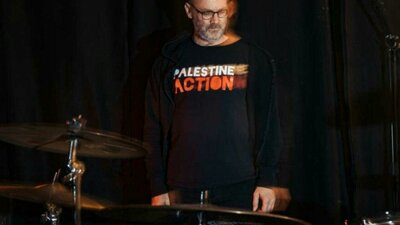 David Rovics standing behind drums with a Palestine Action t-shirt on. Black background.
