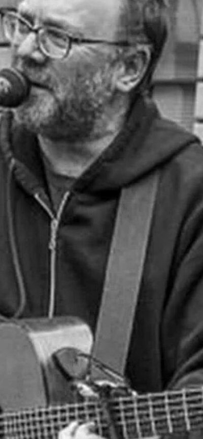 David Rovics in black and white singing and playing the guitar in an outdoor space.