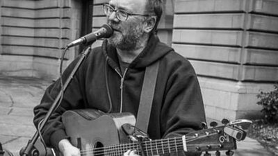David Rovics in black and white singing and playing the guitar in an outdoor space.