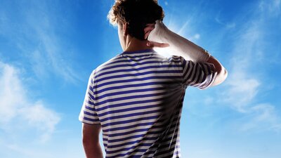 A young man in a blue and white striped t-shit stands facing a blue sky. He has a cast on his arm.