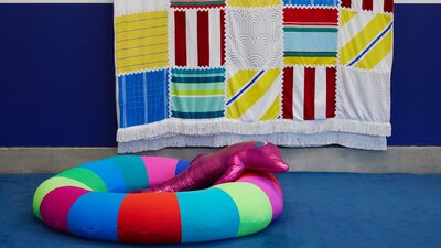 Amy Claire Mills ‘Deep End’, 2025. Liverpool Biennial 2025 at Bluecoat. Pink plush dolphin on an inflatable pool ring and large scale towel hanging in the background