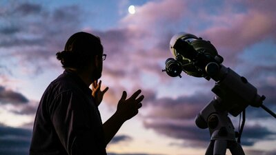 Man looks towards the moon with setting sun next to telescope