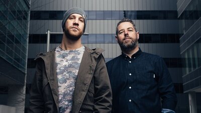Two men standing in front of an office building. One wears a jacket and beanie, the other a shirt