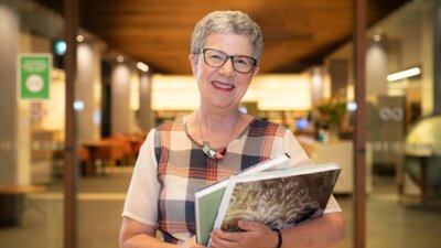 Dr Marie-Louise Ayres FAHA A smiling woman wearing glasses is pictured in a Library holding books
