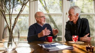 Geoff Pryor and David Pope Two men converse at a wooden table near windows, with a vase of branches and mugs.