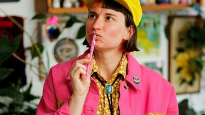 A person with short dark hair and a yellow cap looks thoughtfully upward while holding a pink pen.