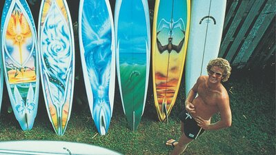 Bombora – The Story of Australian Surfing (two-part series) 2009 Image of man staring up at camera in board shorts, with several surf boards behind him