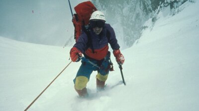A figure in snow gear with snow poles, walking up a snowy mountain towards camera
