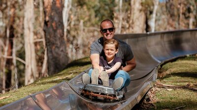 Father and son smiling as they ride on a cart down a metal toboggan track in the forest