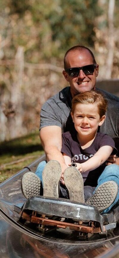 Father and son smiling as they ride on a cart down a metal toboggan track in the forest