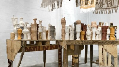 Raw clay vessels on timber tables beneath protest banners in Kara Wood’s Enough installation.
