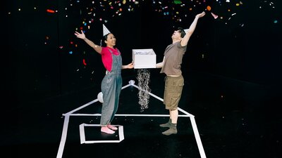 Man and woman holding a gift box and wearing birthday hats throwing colourful confetti into the air