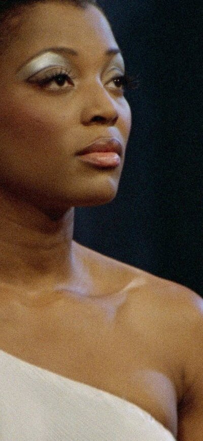 Woman stands with strong eye makeup looking to the right in an off the shoulder white top