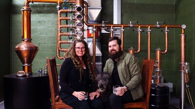 Renee & David (& Parker) are the founders of Everyday Legacy Craft Distillery Renee & David, distillery founders, sitting in front of their still with their dog Parker.