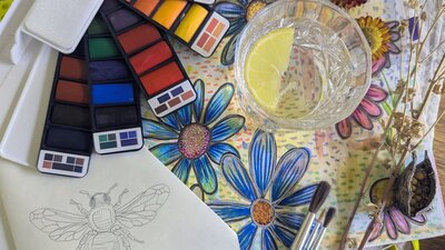 Art workshop samples. Watercolour palletes with painted flowers and a sketched bee next to a gin and tonic.