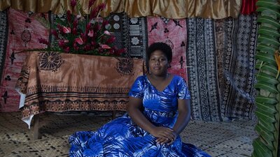 A photograph of a woman sitting down wearing a blue dress in front of multiple layers of fabrics