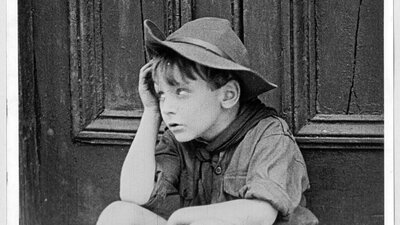 Black and white image of young boy sitting down with a hand on his face rolling his eyes