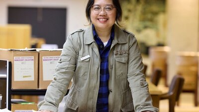 A woman in a green jacket stands leaning on a table covered with papers