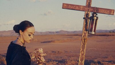 Woman looks down at ground in a dessert with a cross in the ground next to her