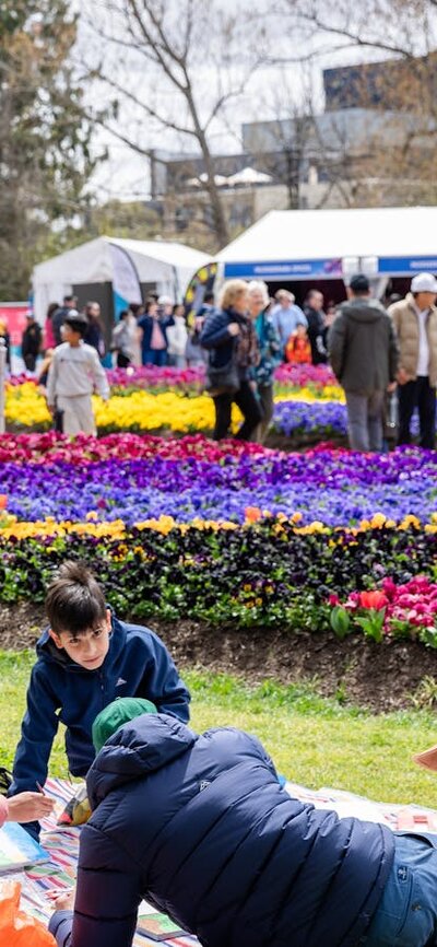 Family picnicking on grass with colourful flower beds and crowds at a flower festival.