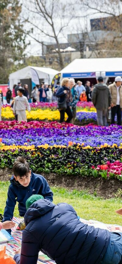 Family picnicking on grass with colourful flower beds and crowds at a flower festival.