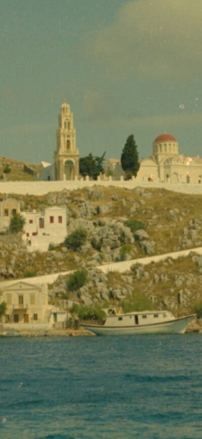 Woman sits on edge of water bank wearing black garment, mountains in background